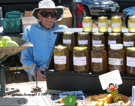 Older gentleman with sunhat and sunglasses with a sales display of mason jars filled with honey in front of him and an old weighting scale filled with green apples to his right.