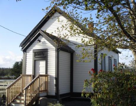 White wooden building with a black gable roof and trimming. The building's entrance has a small wooden stairway. There is a tree and a bush in front right side of the building